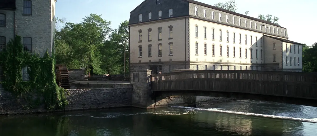 Stone and cream brick mill buildings beside a creek, a wooden covered bridge crossing the water in summer