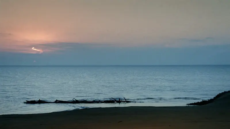 A calm lake at dusk, the last light on the water, a sand beach and driftwood in the foreground