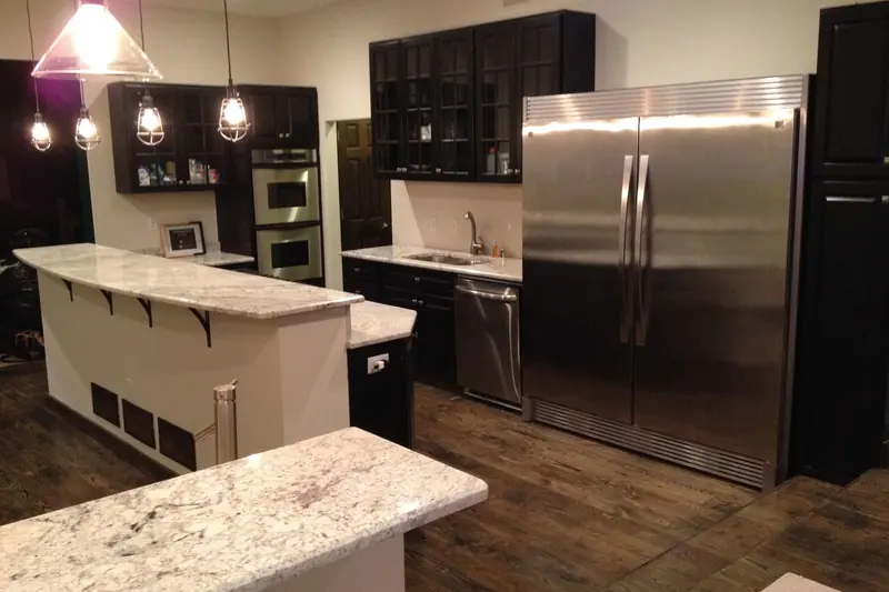 A dark kitchen with a stainless refrigerator, a granite island and pendant lights