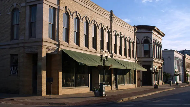A corner of cream brick storefronts with arched windows, green awnings and a gas lamp post in evening light