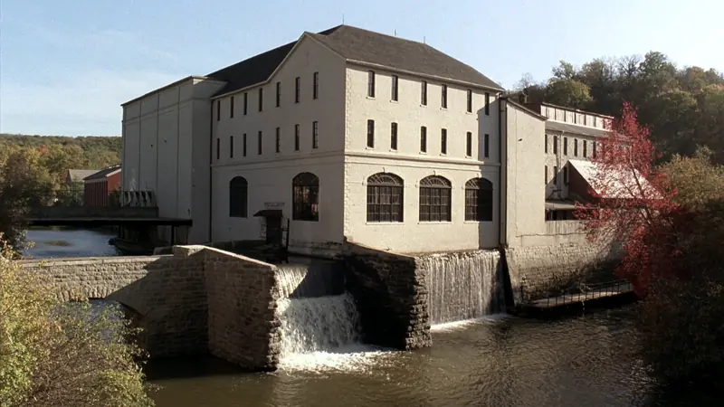 A cream brick mill building above a stone dam, water pouring over the spillway, maples in autumn colour