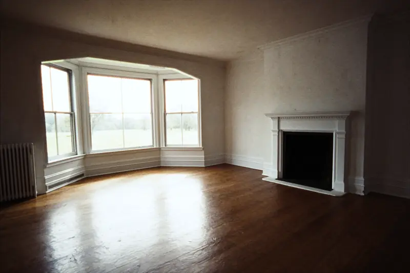 An empty renovated front room with a bay window, a white fireplace mantel and a refinished wood floor